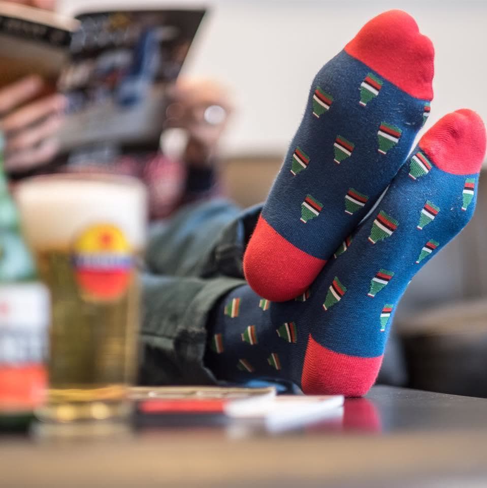 Feet up wearing navy branded socks with a small repeating logo pattern next to a beer