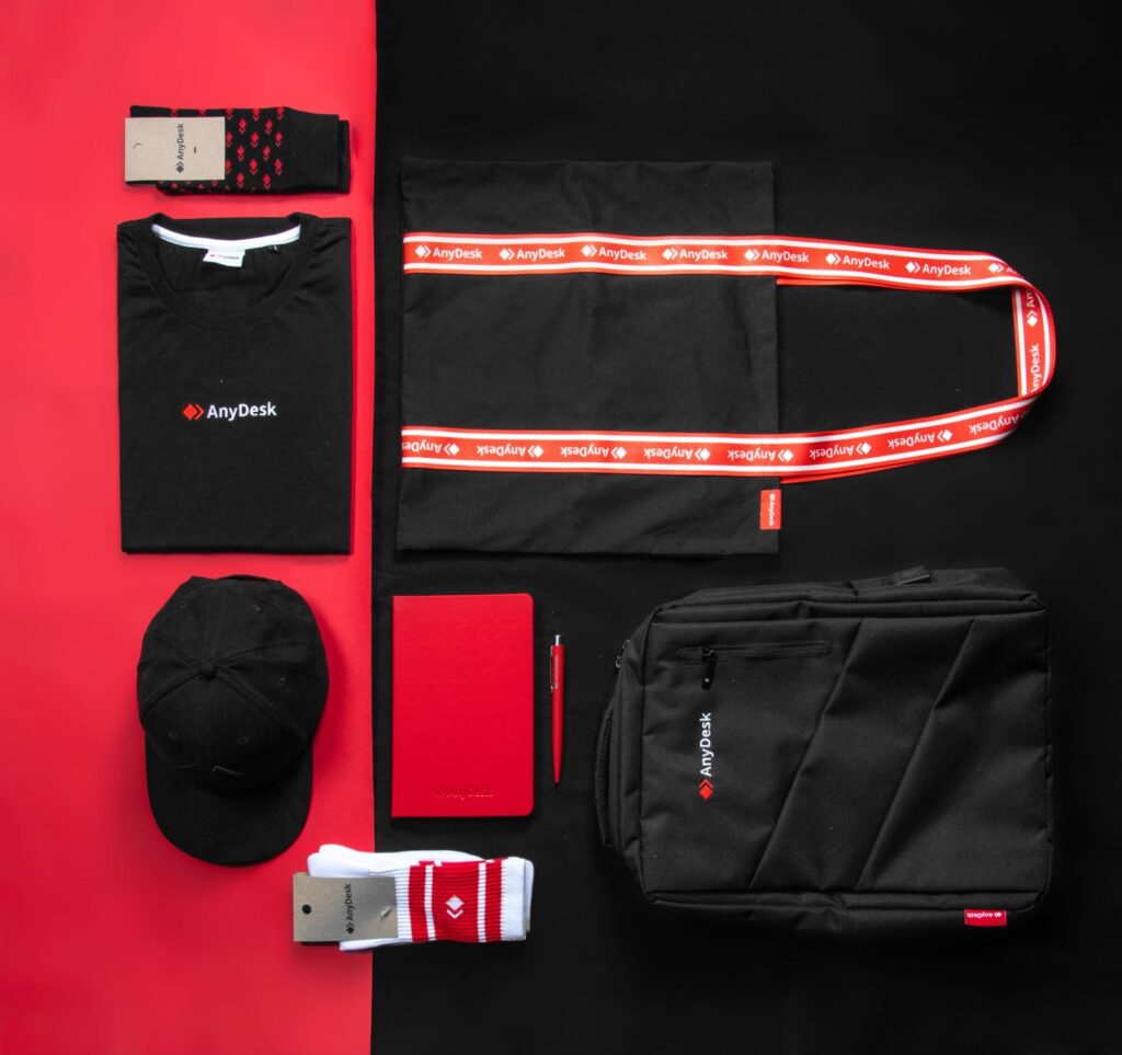 Full AnyDesk merchandise flatlay including t-shirt, cap, socks, notebook, lanyard, tote bag, and backpack